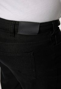 Un pantalon en denim noir présente une étiquette de ceinture en cuir texturé avec "MEN+" en relief. Le pantalon a un design classique à cinq poches.