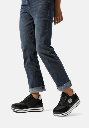 Scarpe sportive nere con una parte superiore testurizzata, suola bianca e dettagli grigi, abbinate a jeans scuri in denim risvoltati.