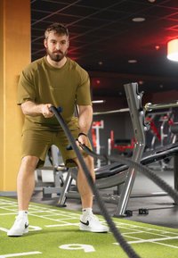 Homme en t-shirt et short de sport vert olive exerçant une force sur de grosses cordes noires dans une salle de sport équipée de matériel gris et rouge. Il porte des baskets blanches sur un gazon synthétique vert.