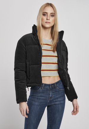 Svart kordfløyelsjakke i cropped design med glidelås foran og høy krage. Gråstripete skjorte under. Modellen har på seg mørkeblå jeans.