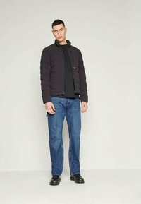 Casaco preto acolchoado com fecho à frente, combinado com uma camisa preta e jeans azuis. Modelo usa sapatos pretos. Design simples, sem padrões visíveis.