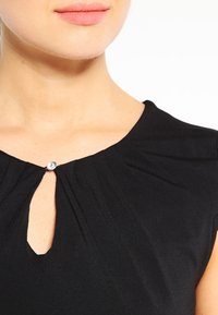 Dolna część twarzy i szyja kobiety w czarnej bluzce z małym otworem w kształcie kropli i jednym przezroczystym guzikiem przy dekolcie.