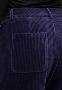 Giluoti tamsiai purpuriniai kordūrai, su vertikaliu raštu, turintys galinį kišenę ir pritaikytą juosmenį. Tekstūra atrodo švelni ir aksominė.