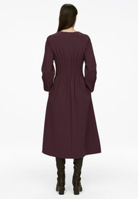 Langärmliges Kleid in Dunkellila mit einem gepleiteten Rückendesign. Hergestellt aus glattem Stoff, mit rundem Ausschnitt und knöchellangem Schnitt.