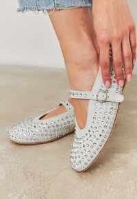 Kobieta w jasnoszarych zamszowych butach Mary Jane z metalowymi ćwiekami i paskami na klamry, poprawiająca jeden but na beżowym tle.