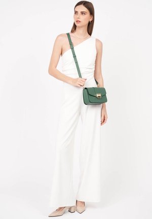 Weißer Jumpsuit mit einem Schulterdesign, weitem Bein; Model hält eine grüne Umhängetasche mit einem goldenen Verschluss; beige Pumps mit spitzer Zehen.