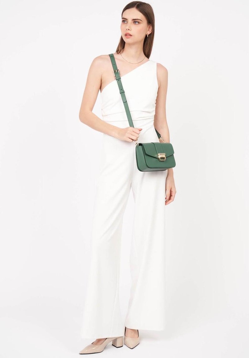 Weißer Jumpsuit mit einem Schulterdesign, weitem Bein; Model hält eine grüne Umhängetasche mit einem goldenen Verschluss; beige Pumps mit spitzer Zehen.