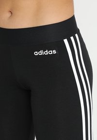 Czarne legginsy sportowe z szerokim pasem, z trzema białymi paskami po bokach i małym białym logo adidas.