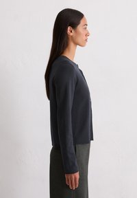 Sieviete ar garām tumšām matēm, tērpusies ar pogātu tumšu kardiganu un pelēkiem svārkiem, stāv profilā pret vienkāršu baltu fonu.