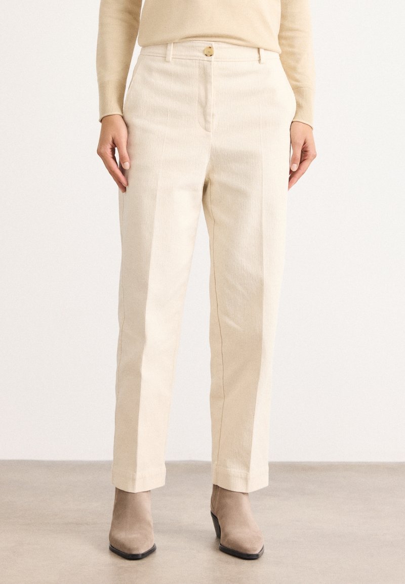 Femme portant un pantalon en velours côtelé beige, un pull beige assorti et des bottines en daim taupe, debout sur un sol gris.