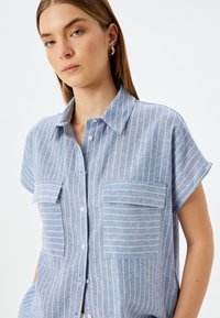 Camisa de manga corta azul claro con rayas verticales blancas, dos bolsillos en la parte frontal y cierre de botones. Textura suave y diseño de cuello.