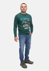 Maglione verde con motivi "St. Barth" e per lo sci, jeans blu e scarpe sportive grigie. Il maglione presenta grafiche ricamate e un colletto rotondo.