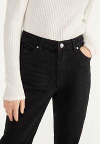 Vaqueros de mezclilla negros de cintura alta, con cinco bolsillos y cierre de botón plateado. Combinados con una camiseta blanca de manga larga acanalada. Textura suave.