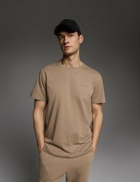 Beżowa bawełniana koszulka z krótkimi rękawami i okrągłym dekoltem, noszona z dopasowanymi beżowymi spodniami; czarna czapka dopełnia całość.