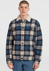 Young man wearing a blue and white plaid jacket with black buttons and a cream-colored fleece collar, paired with blue pants.