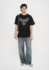 Svart oversized T-skjorte med ørn-grafikk, paret med løse blå jeans og svart-hvite sneakers. Tilbehør inkluderer en sølvkjede-veske.