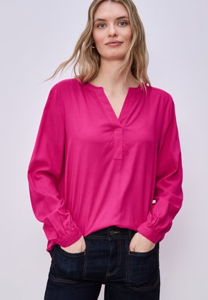 Kobieta o jasnych włosach, ubrana w jaskraworóżową bluzkę z długim rękawem i ciemnoniebieskie dżinsy, stojąca z rękami w kieszeniach na jednolitym tle.