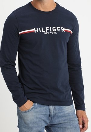 Marineblaues Langarm-T-Shirt mit dem Aufdruck "HILFIGER NEW YORK" in Weiß, akzentuiert mit roten und weißen Streifen. Baumwollmaterial, Rundhalsausschnitt.