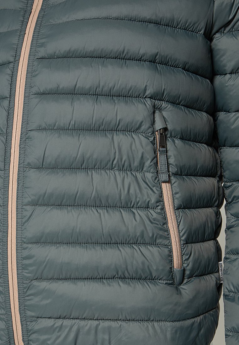 Veste verte rembourrée avec un design matelassé vertical, fermeture zippée à l'avant et une poche latérale. Comprend un zip contrastant.