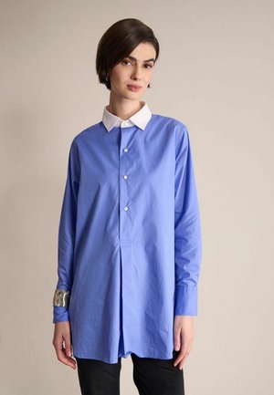 Femme aux cheveux courts et sombres portant une longue chemise bleue à col blanc, un bracelet manchette argenté et un pantalon noir sur un fond uni.