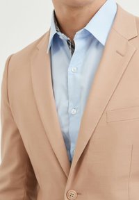 Blazer beige avec une texture lisse et une coupe ajustée, avec une chemise à manches longues bleu clair en dessous. Motif décoratif visible à l'intérieur du col.
