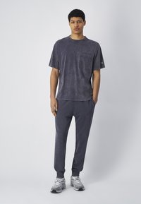 Camiseta de algodón de color gris oscuro, ligeramente texturizada, con un bolsillo en el pecho, combinada con pantalones jogger a juego que presentan un logo grabado y piernas ajustadas.