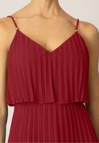 Femme portant une robe sans manches, plissée, rouge foncé avec des bretelles fines et des accents en perles argentées sur les épaules.