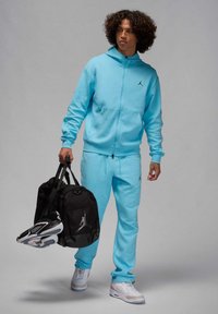 Completo felpa e pantaloni da jogging azzurro chiaro con dettagli logo neri, abbinato a una borsa nera e sneakers con dettagli bianchi e grigi.