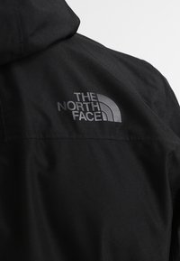 Schwarze Jacke mit Kapuze, strukturiertem Stoff und einem grauen Logo von "The North Face" auf dem oberen Rücken. Schlichtes Design, keine Muster.