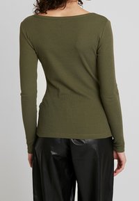 Langärmliges, olivgrünes Oberteil aus Rippstoff. Verfügt über einen weiten Ausschnitt und eine taillierte Silhouette, kombiniert mit schwarzen, glänzenden, weit geschnittenen Hosen.