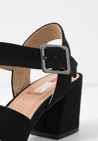 Sandale à talon bloc en daim noir avec une bride à la cheville et une boucle carrée en argent, semelle intérieure beige lisse et rembourrage en mousse douce.