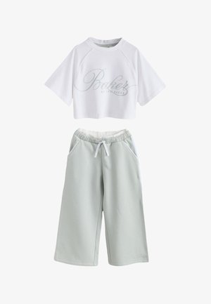 Młoda kobieta z długimi ciemnymi włosami patrzy na bok, nosząc biały sweter z wyszywanym logo "Baker by Ted Baker".