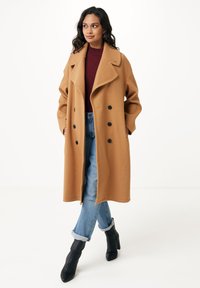 Cappotto cammello doppiopetto con ampi revers, bottoni neri e tasche laterali, abbinato a un maglioncino bordeaux e jeans blu con risvolto. Stivali neri alla caviglia.