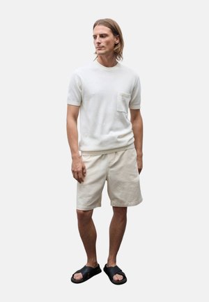 Homme debout portant un pull en maille écru à manches courtes avec une poche poitrine, un short beige et des sandales noires à bout ouvert.