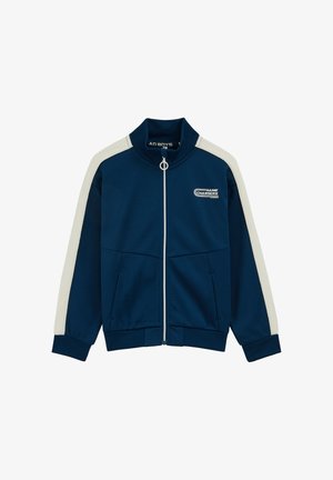 Temno modra jopa z zadrgo, belimi črtami na rokavih, visokim ovratnikom, sprednjimi žepi in logotipom »Game Changers« na levi strani prsnega koša.