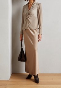 Blouse beige à boutons avec un détail noué à la taille, associée à une jupe beige satinée longueur cheville. Talons aiguilles noirs et un sac noir.