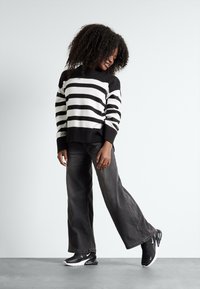 Pullover a righe bianche e nere con orlo a costine, abbinato a jeans grigi a gamba ampia e scarpe sportive nere con accenti bianchi.