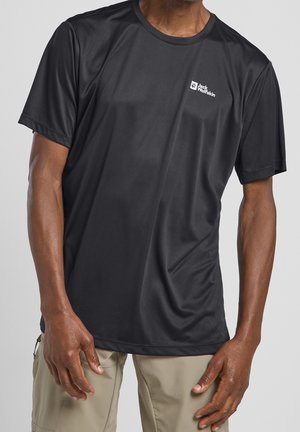 Man wearing black short-sleeve Jack Wolfskin athletic shirt and beige pants, standing against a plain light gray background.