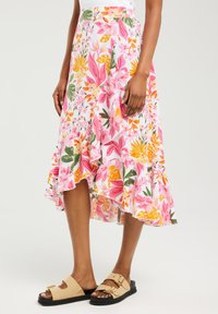 Jupe maxi florale avec un fond rose, ornée de motifs jaunes et verts. Ourlet à volants et tissu léger. Sandales beige avec boucles.