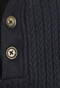 Donkergrijze gebreide stof met gevlochten patroon en twee grote, ronde, glanzende knopen genaaid met zwarte draad.