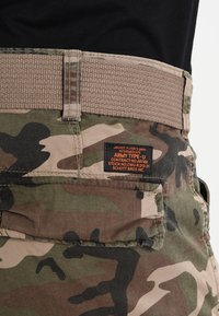 Tarnfarben Cargo-Hosen mit einem strukturierten beigen Bund und einem schwarzen Etikett, das militärische Spezifikationen und Branddetails anzeigt.