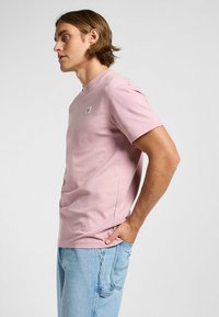 Lyserød t-shirt med korte ærmer lavet af bomuld, med en rund halsudskæring og et lille logo-badge på brystet. Båret med lyseblå jeans.