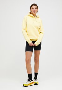 Gelbes Hoodie mit einer Fronttasche und Kordelzug, kombiniert mit schwarzen Fahrradhosen und mehrfarbigen Sneakers. Das Modell steht vor einem einfarbigen Hintergrund.