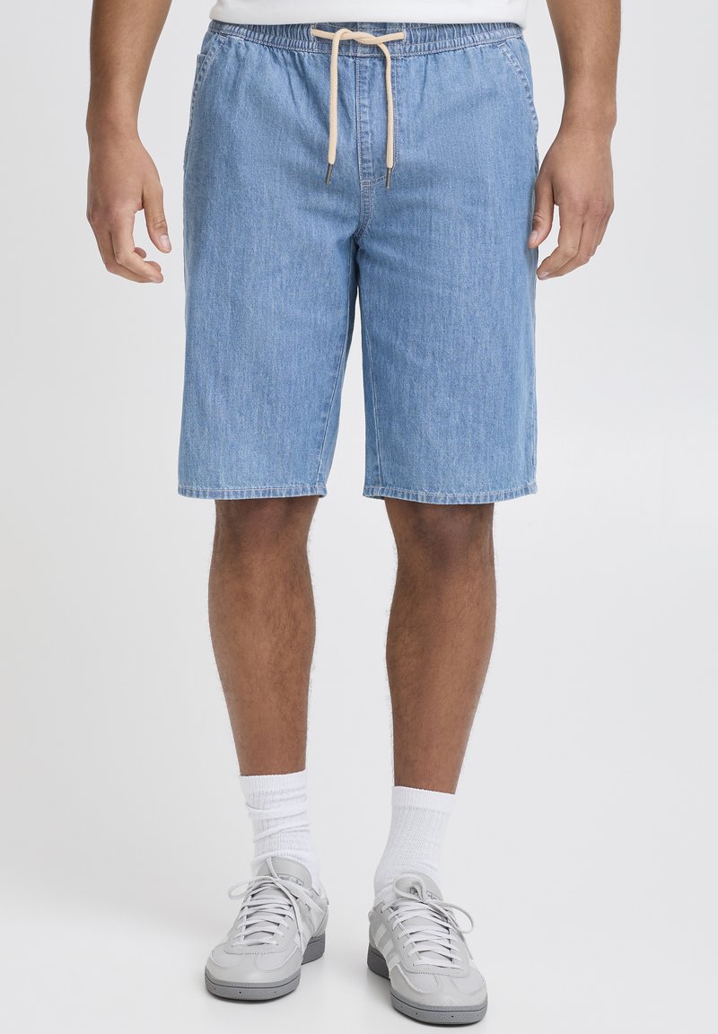 Shorts di denim azzurro chiaro con vita regolabile a coulisse, caratterizzati da una vestibilità rilassata e lunghezza fino al ginocchio. Indossati con calzini bianchi e scarpe da ginnastica grigie.