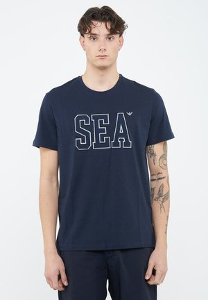 Jonge man met donker golvend haar, gekleed in een marineblauw SEA T-shirt en zwarte broek, zichtbare tatoeages op de rechteronderarm, staand tegen een effen witte achtergrond.