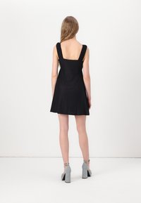 Vestido preto em linha A com decote quadrado e alças largas. O tecido tem um padrão texturizado. Combinado com sapatos de salto alto cinzas.