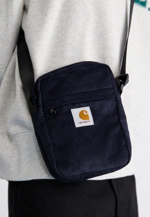 Kleine dunkelblaue Cord-Schultertasche mit Carhartt-Logo-Patch, getragen über einem grauen Sweatshirt und schwarzen Hosen.