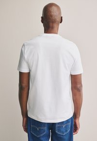 Maglietta bianca in cotone a maniche corte, collo rotondo e taglio semplice; abbinata a jeans blu in denim con tasche posteriori e dettagli di cucitura.