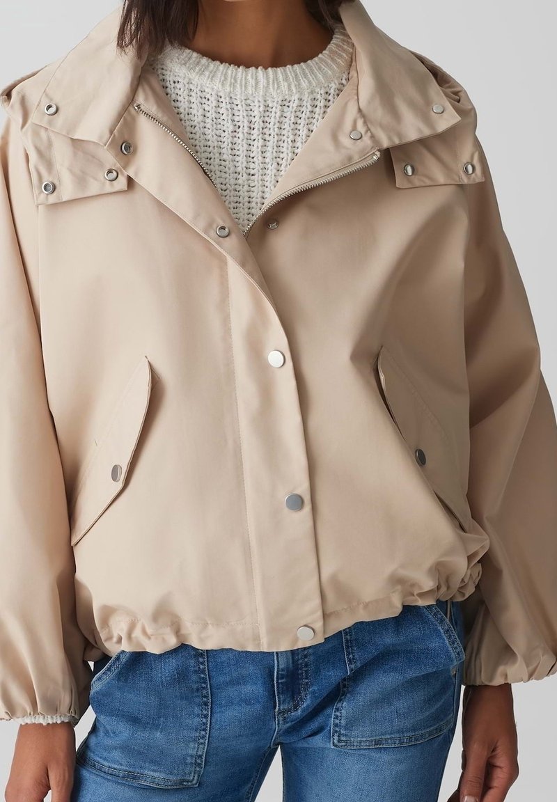 Giacca beige leggera con bottoni a pressione, orlo elasticizzato, due tasche laterali e colletto pieghevole. Indossata sopra un maglione bianco in maglia.
