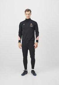 Musta urheilullinen harjoitusasu vaaleanpunaisilla yksityiskohdilla, vetoketjullinen takki ja slim-fit-housut, joissa on logoja, sekä mustat urheilukengät sinisillä yksityiskohdilla.